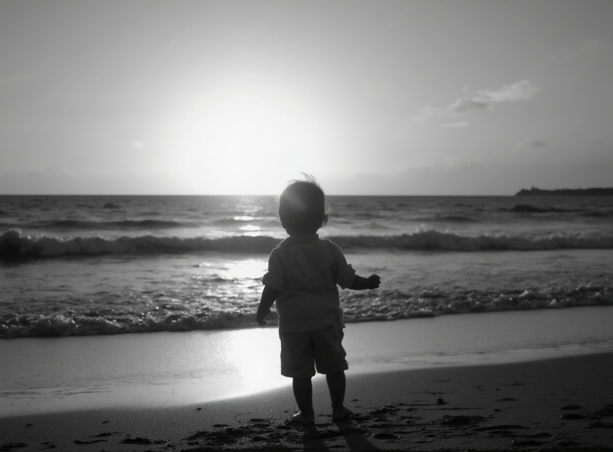 A black-and-white photograph of a child at the beach at sunset. The child is looking out toward the setting sun.