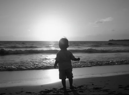 A black-and-white photograph of a child at the beach at sunset. The child is looking out toward the setting sun.