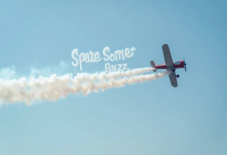A biplane during air show after which the condensation trail is shaped in the text "spare some buzz". Text "spare some buzz" is on the sky