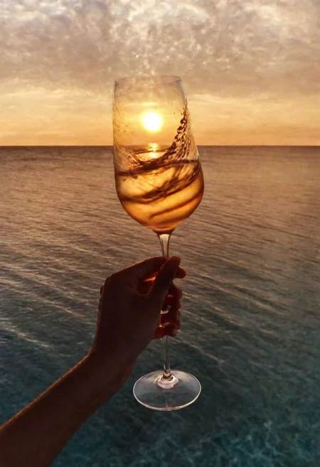 F-B-C, POV, foreground: a hand stir a glass filled with water   , background: sunset on the sea , effect: sunset reflecting into the liquid filling the glass