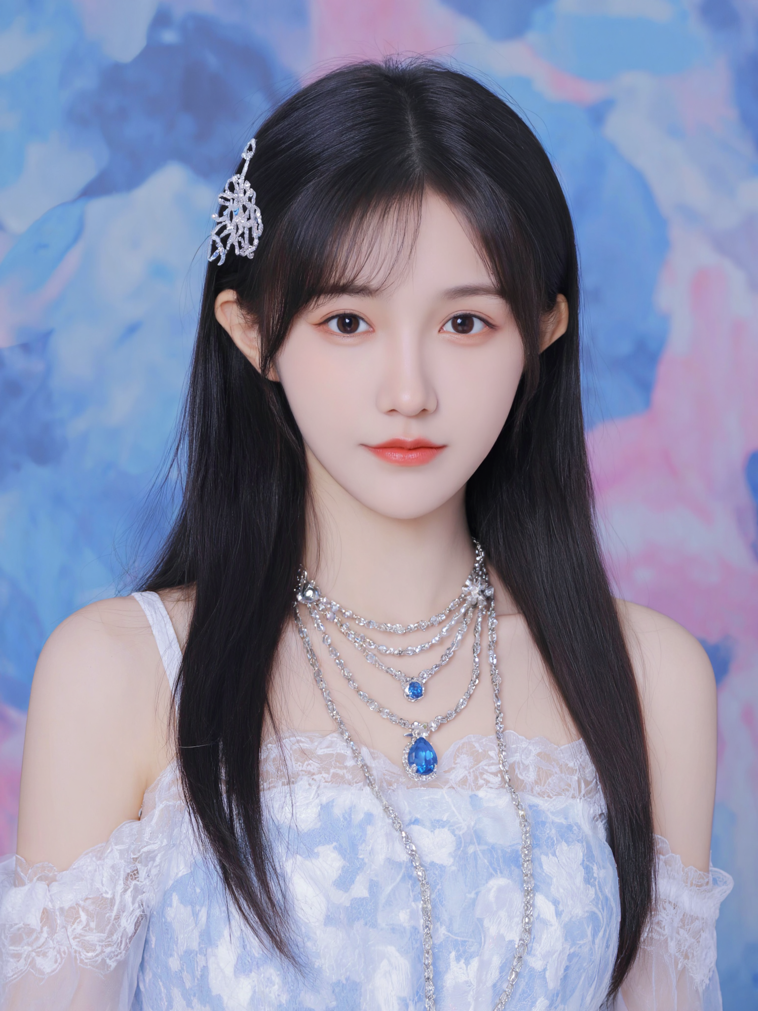 This is a high-resolution photograph featuring a young Asian woman with fair skin and long, straight black hair adorned with a delicate, white floral hair clip. She wears an intricate, light blue and white lace dress with detailed embroidery, and multiple layers of blue and clear gemstone necklaces. The background is an abstract, colorful blend of blue and pink hues, creating a vibrant contrast to her pale skin and elegant attire. The lighting is soft and even, highlighting her serene expression and the texture of her dress. The camera angle is slightly low, capturing her from the waist up, and the composition follows the rule of thirds, drawing attention to her face and jewelry.
