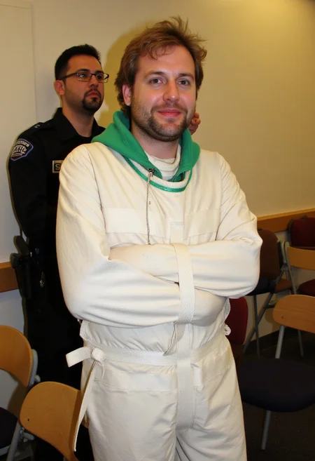 A photograph, likely in a lecture room. In the background, a man with short black hair and a beard, wearing a security guard uniform and glasses, is standing and doing something behind second person. The second person, a middle aged man with light brown wavy hair, untidy beard and earring is wearing a white sjposey, with a green hood behind his neck. He has a slight smile, suggesting a nervous mood. The room features a mix of wooden and upholstered chairs, adding to the casual atmosphere. <lora:straitjacketflux-posey:1.1>
