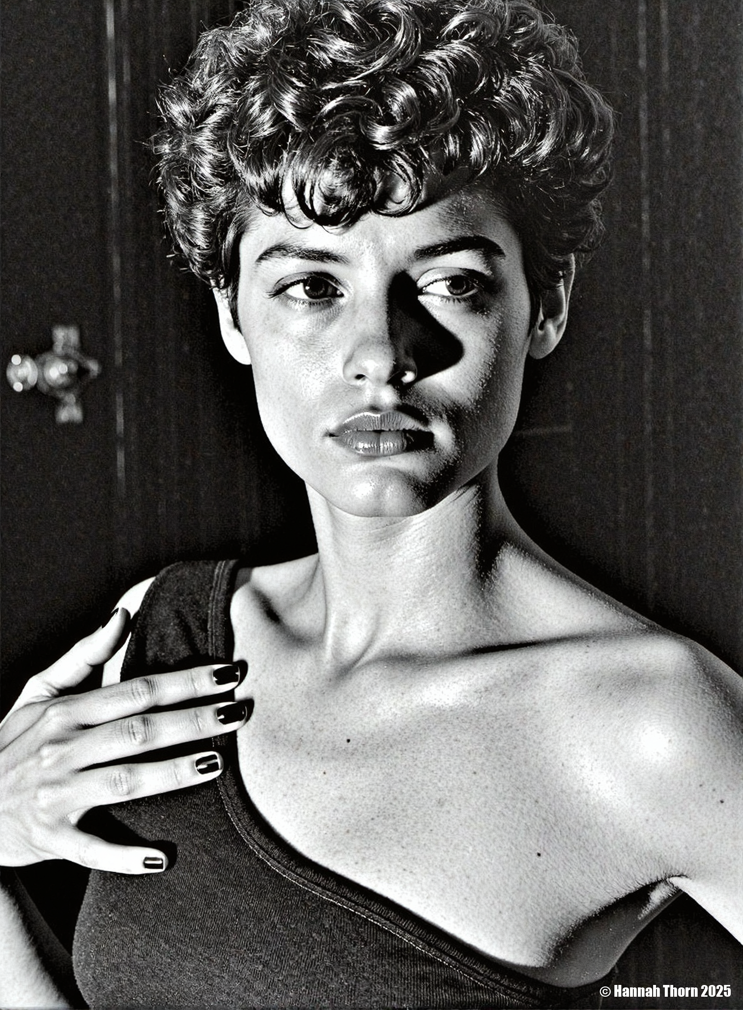 Generate a high-contrast, black and white portrait photograph, reminiscent of Helmut Newton and Peter Lindbergh, depicting a young woman from the chest up. Her short, curly hair frames her face, subtly angled away from the camera towards her right shoulder. She wears a dark, sleeveless top with a visible neckline, her right hand resting near her shoulder, partially obscuring the garment. Employ chiaroscuro lighting with strong shadows, creating a shallow depth of field that emphasizes the woman's face, leaving the dark, indistinct background softly blurred.  The image should exhibit a grainy film aesthetic, recalling the textures of classic silver halide photography, with a monochrome palette intensified by strong tonal separation à la Irving Penn.  Infuse the style with a touch of the dramatic intensity found in the works of Edward Hopper, and ensure a sense of mystery and quietude in the subject's expression, achieving a final image with high resolution and rich detail. <lora:_Flux\_Photographer\Helmut_Newton-000001.safetensors:0.85:0.85> <lora:_Flux\_Skin_Appearance\Detailed_Breasts-000004.safetensors:0.75:0.75>